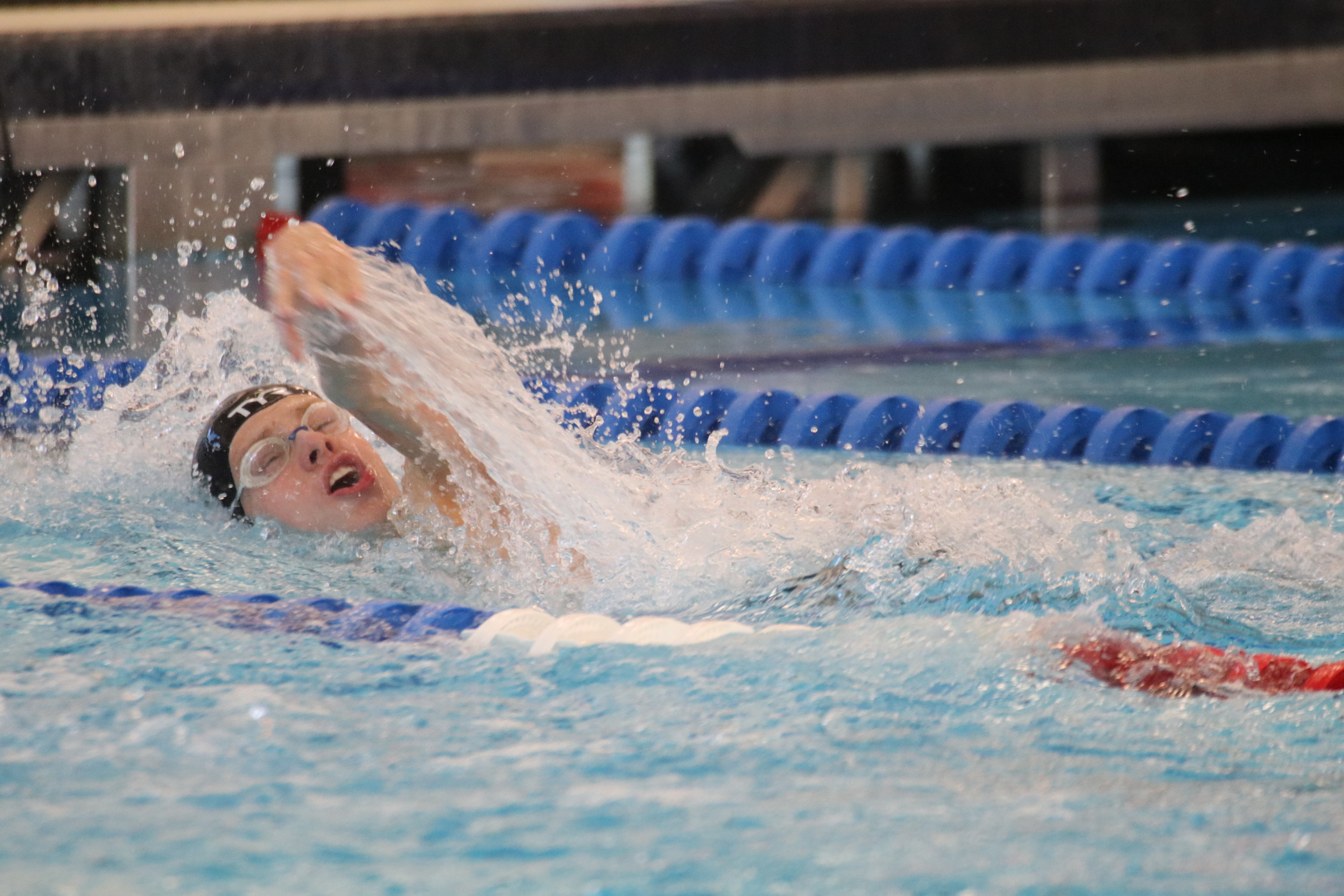 A swimmer wearing a black TYR cap and goggles performs a freestyle stroke, his arm mid-recovery above a splash of water in a competitive pool with blue lane lines.