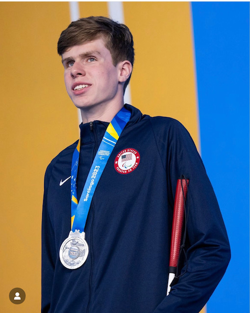 A young man in a navy blue Team USA jacket looks upward with a smile while wearing a silver Santiago 2023 medal around his neck. He is holding a red and white mobility cane.