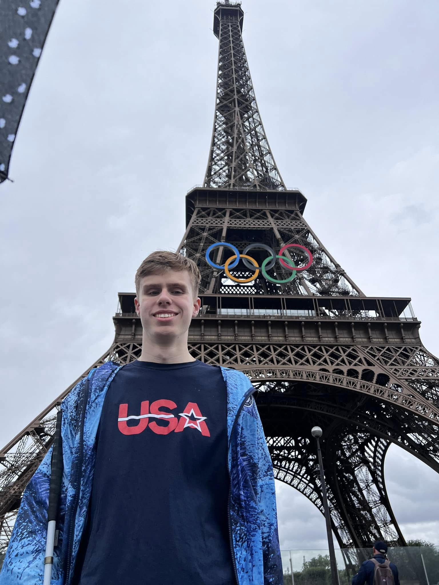 A smiling young man stands in front of the Eiffel Tower in Paris, which is adorned with the Olympic rings. He is wearing a navy blue USA t-shirt and a blue patterned zip-up jacket, holding a white mobility cane in his right hand.