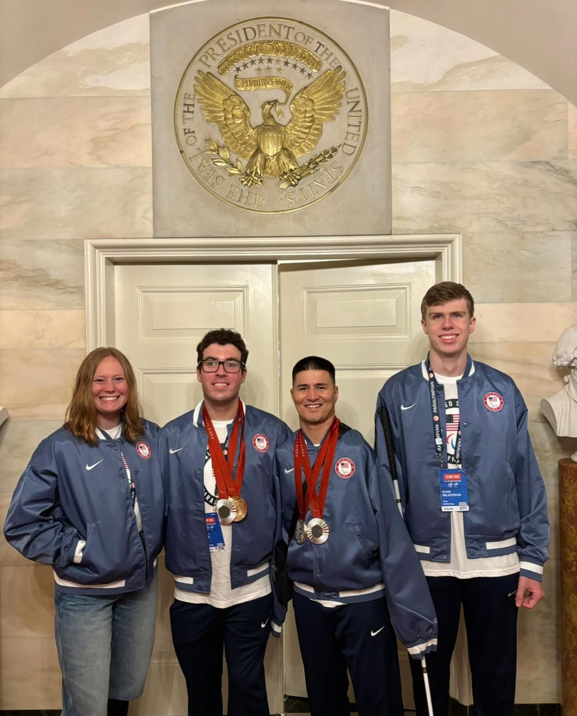 Four Team USA athletes in matching blue jackets stand together for a photo in a formal room under a large gold Seal of the President of the United States. Two of the athletes are wearing medals.