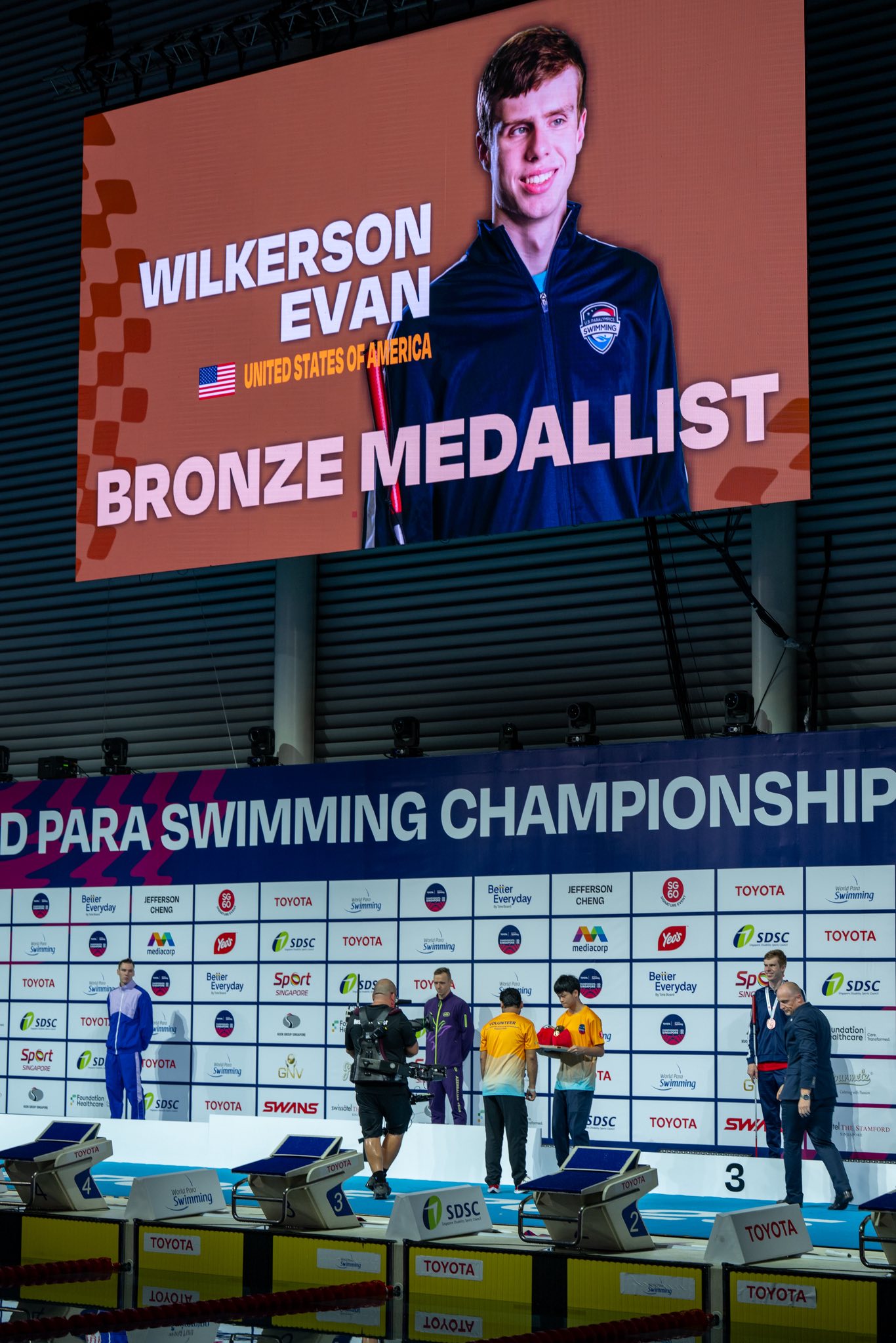 A wide shot of a World Para Swimming Championship arena. A massive screen displays a portrait of Evan Wilkerson with the text BRONZE alongside competition information.